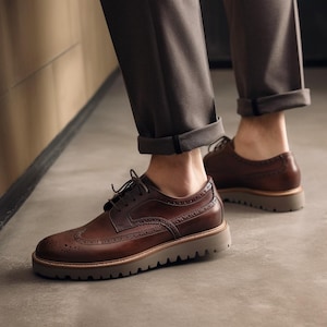 Peut inclure: Chaussures brogues en cuir marron avec une semelle épaisse marron clair. Les chaussures présentent des perforations décoratives et sont lacées. L'image montre les chaussures sous un angle bas, mettant en valeur leur design. Elles sont portées avec un pantalon gris.