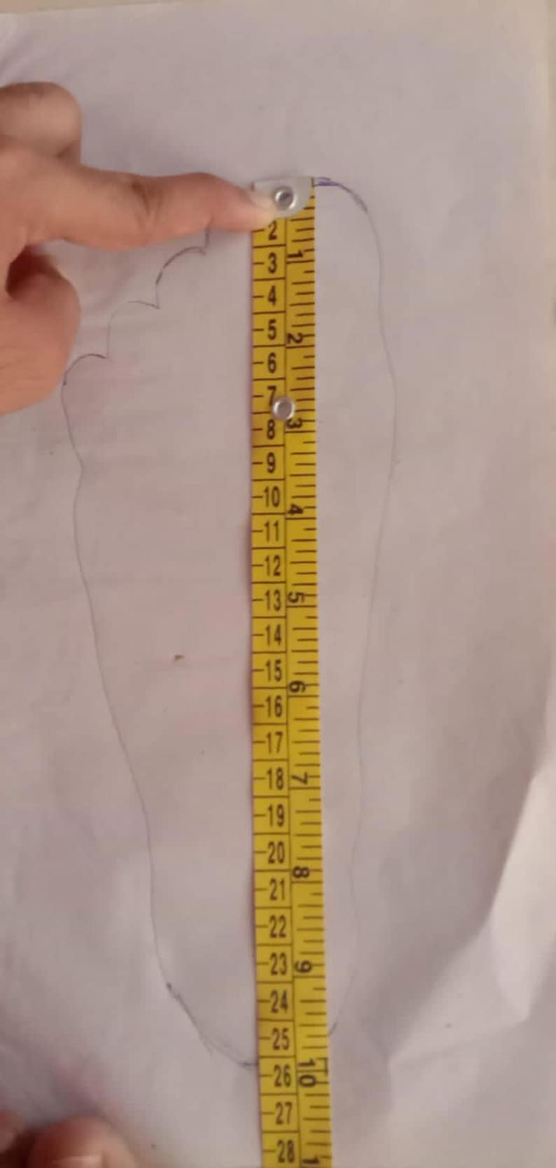 May include: A yellow measuring tape is placed next to a traced outline of a foot on white paper. The tape shows measurements in inches, with the foot outline measuring approximately 10 inches in length. The image is taken from above.