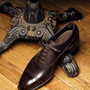 May include: A brown leather dress shoe with a cap toe and lace-up closure. The shoe is resting on a wooden surface near a decorative metal object and a table leg.