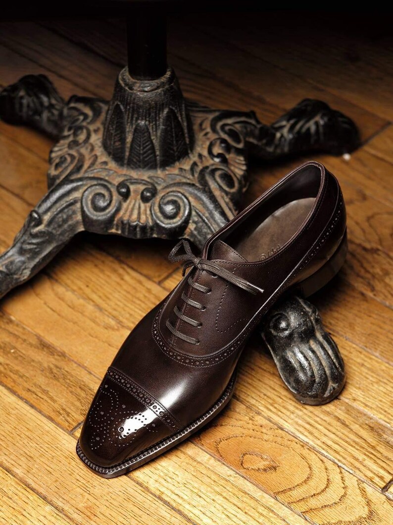 May include: A brown leather dress shoe with a cap toe and lace-up closure. The shoe is resting on a wooden surface near a decorative metal object and a table leg.