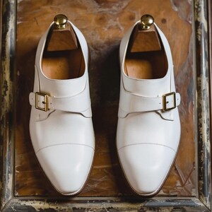 May include: A pair of white leather monk strap shoes with gold-colored buckles. The shoes are displayed on wooden shoe trees, showcasing their classic design and craftsmanship. The shoes have a pointed toe and a strap across the top.