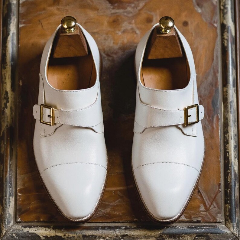 May include: A pair of white leather monk strap shoes with gold-colored buckles. The shoes are displayed on wooden shoe trees, showcasing their classic design and craftsmanship. The shoes have a pointed toe and a strap across the top.