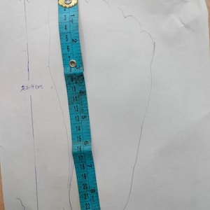 Op de afbeelding: Een blauwe meetlint ligt naast een getraceerde voetomtrek op wit papier. De voetomtrek meet 23,4 cm in lengte en 10,5 cm in breedte. De tape toont metingen in centimeters.
