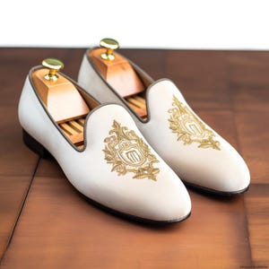 Op de afbeelding: Een paar cr&egrave;mekleurige loafers met goud geborduurde wapenschilden. De schoenen hebben een ronde neus, een zwarte zool en een grijze bies. Houten schoenspanners zitten in de schoenen. De loafers worden op een houten oppervlak getoond.
