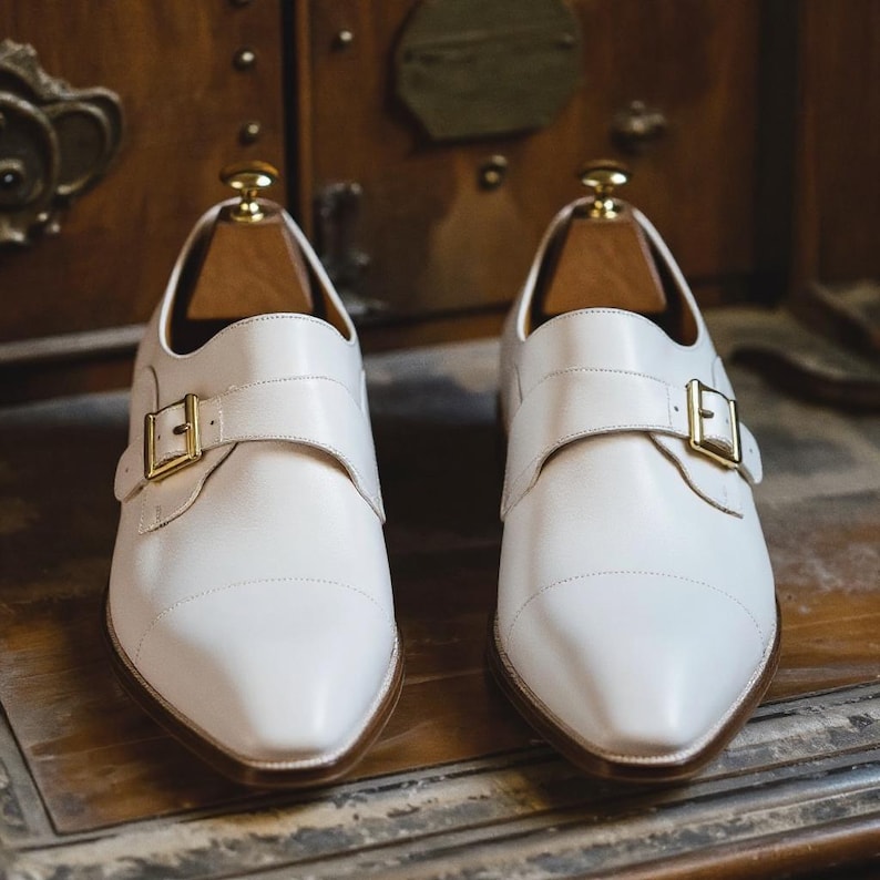 May include: A pair of white leather dress shoes with a buckle closure and wooden shoe trees. The shoes have a pointed toe and a gold-colored buckle. The shoes are displayed on a wooden surface.