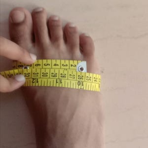 May include: A person's foot being measured with a yellow measuring tape. The tape is wrapped around the foot, showing the measurement in inches. The image focuses on the foot and the measuring process.