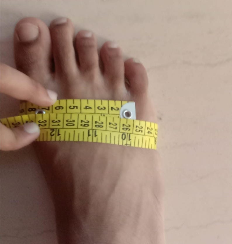 May include: A person's foot being measured with a yellow measuring tape. The tape is wrapped around the foot, showing the measurement in inches. The image focuses on the foot and the measuring process.