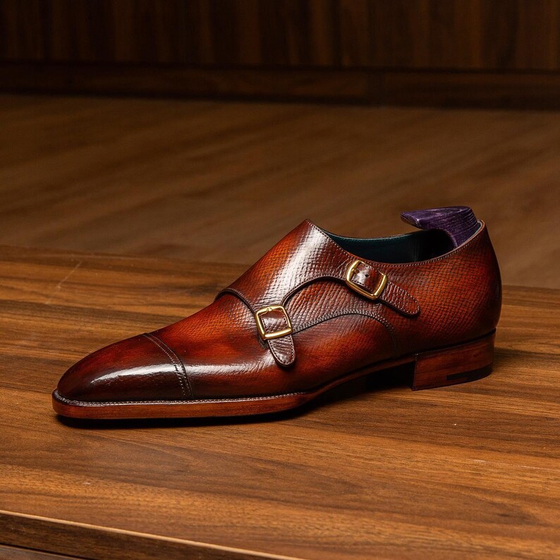 May include: A brown leather double monk strap shoe with a textured finish. The shoe features two gold-colored buckles and a pointed toe. The shoe is displayed on a wooden surface.