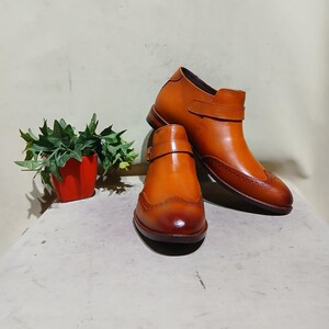 May include: A pair of brown leather ankle boots with decorative brogue detailing and buckle closures. The boots have a polished finish and are set against a neutral background. A small red pot with green plants is in the background.