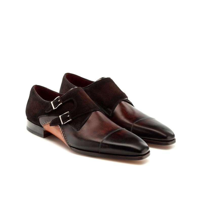 May include: A pair of brown leather dress shoes with a cap toe design. The shoes feature a double monk strap closure with silver buckles and a suede accent. The shoes are a classic style.