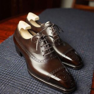 May include: A pair of brown leather dress shoes with a glossy finish. The shoes feature a classic Oxford design with intricate brogue detailing and lace-up closures. Wooden shoe trees are inserted inside the shoes.