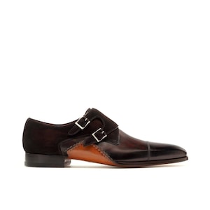 May include: A brown leather double monk strap shoe with a dark brown suede accent. The shoe features two silver buckles and a stitched design along the side. The toe is a darker shade of brown.