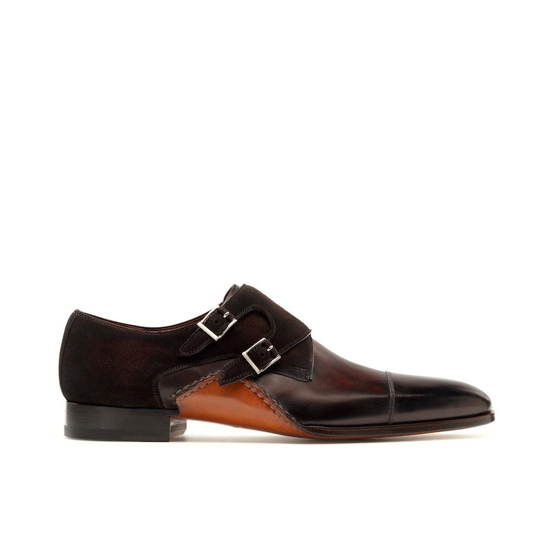 May include: A brown leather double monk strap shoe with a dark brown suede accent. The shoe features two silver buckles and a stitched design along the side. The toe is a darker shade of brown.