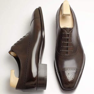 May include: Two brown leather dress shoes with wooden shoe trees inside, set against a white background. The shoes have a polished finish, decorative perforations, and a classic lace-up design. The shoes are a classic style.