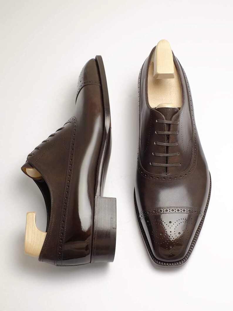 May include: Two brown leather dress shoes with wooden shoe trees inside, set against a white background. The shoes have a polished finish, decorative perforations, and a classic lace-up design. The shoes are a classic style.