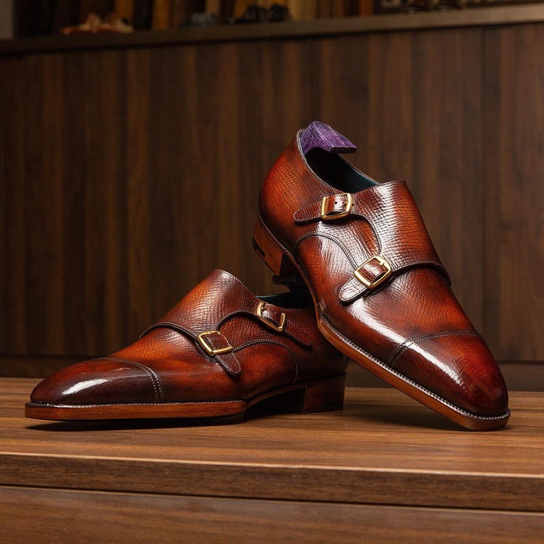 May include: A pair of brown leather double monk strap shoes with gold-colored buckles. The shoes have a textured finish and a polished appearance, with a dark brown toe cap. The interior of the shoe is purple. The shoes are displayed on a wooden surface.