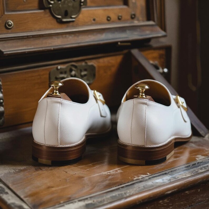 May include: A pair of white leather dress shoes with gold-colored buckles and wooden shoe trees. The shoes have brown soles and are displayed on a wooden surface. The background includes a wooden cabinet.