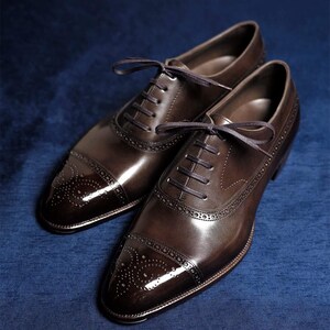 May include: A pair of brown leather Oxford shoes with a glossy finish. The shoes feature a cap toe design with decorative perforations and dark brown laces. The shoes are displayed on a blue surface.