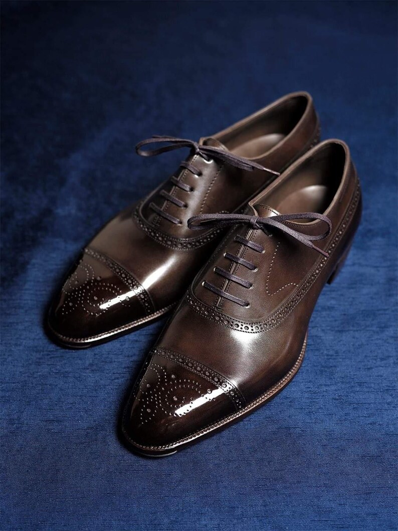 May include: A pair of brown leather Oxford shoes with a glossy finish. The shoes feature a cap toe design with decorative perforations and dark brown laces. The shoes are displayed on a blue surface.