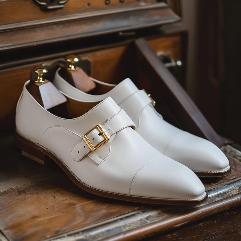 May include: A pair of white leather monk strap shoes with gold-colored buckles. The shoes have a brown sole and are displayed on a wooden surface. The shoes are new and unworn.