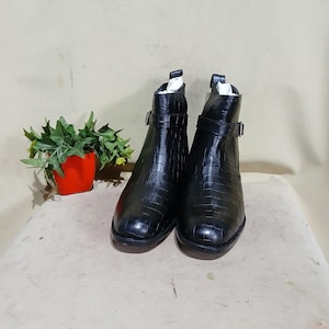 Peut inclure: Paire de bottines noires avec une texture peau de crocodile. Chaque botte a une bride à boucle et un petit talon. Les bottes sont présentées sur une surface neutre, avec une petite plante en pot en arrière-plan.