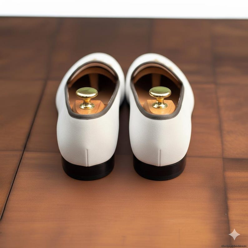 Op de afbeelding: Een paar witte leren loafers met donkere zolen en grijze rand. Elke schoen heeft een houten schoenspanner met een goudkleurige knop. De schoenen worden gepresenteerd op een bruin houten oppervlak.
