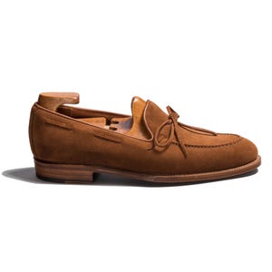 May include: A brown suede loafer with a leather sole and a small heel. The shoe features a decorative bow detail on the vamp and a wooden shoe tree inside. The overall design is classic and elegant.