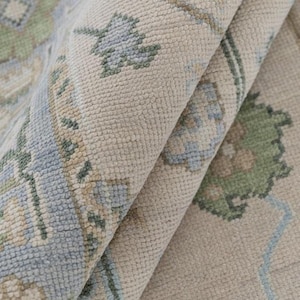 Puede incluir: Primer plano de una alfombra doblada con un patrón floral detallado. La alfombra presenta un fondo beige con diseños florales azules y verdes. La textura de la alfombra es visible, mostrando una construcción tejida.