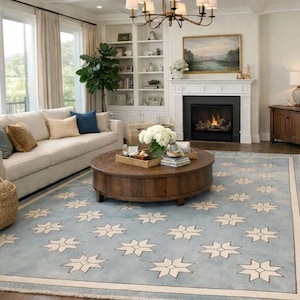 Könnte beinhalten: Wohnzimmer mit hellblauem Teppich mit sich wiederholendem Sternmuster. Ein runder Couchtisch aus Holz steht in der Mitte, umgeben von einem beigen Sofa, einem Kamin und einem Kronleuchter. Der Raum ist in neutralen Tönen gehalten.