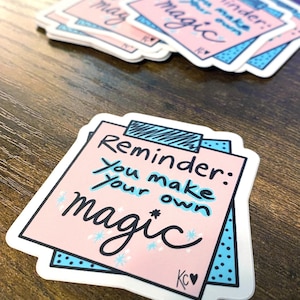 May include: Pink and blue stickers with the text "Reminder: You make your own magic." The stickers have a white border and a blue dotted pattern. The stickers are stacked on a wooden surface.