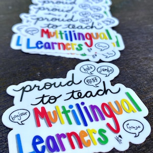 May include: White stickers with the text "proud to teach Multilingual Learners" in a colorful design. The word "Multilingual" is in rainbow colors. Speech bubbles with greetings in different languages are included.