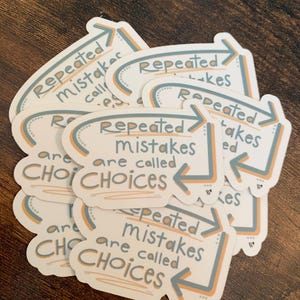 K&ouml;nnte beinhalten: Ein Stapel wei&szlig;er Aufkleber mit dem Text "Repeated mistakes are called choices" in handschriftlicher Schrift. Die Aufkleber haben ein hellblaues und orangefarbenes Pfeildesign.