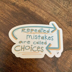 K&ouml;nnte beinhalten: Ein wei&szlig;er Aufkleber mit dem Text "Repeated mistakes are called CHOICES" in T&uuml;rkis und Orange. Der Text befindet sich in einer wei&szlig;en Form mit t&uuml;rkisfarbenen und orangefarbenen Pfeilen und Akzenten. Der Aufkleber befindet sich auf einer braunen Holzoberfl&auml;che.