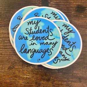 May include: A stack of round stickers with a blue and green world map design. The stickers have the text "my students are loved in many languages" in black cursive font.