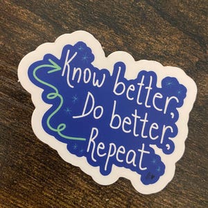 May include: A white sticker with the text "Know better, Do better, Repeat" in a handwritten style. The text is on a blue background with a green arrow and swirls. The sticker is on a brown wooden surface.
