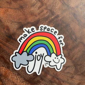 May include: A colorful sticker featuring a rainbow with the words "make space for joy." The rainbow has red, orange, yellow, green, and blue arches. Gray clouds flank the rainbow, and a smiling cloud is on the right.