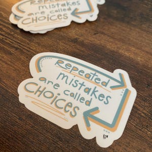 K&ouml;nnte beinhalten: Wei&szlig;er Aufkleber mit dem Text "Repeated mistakes are called CHOICES" in einer verspielten Schriftart. Der Text ist von einem hellblauen und orangefarbenen Design umgeben. Mehrere Aufkleber sind auf einer Holzoberfl&auml;che zu sehen.