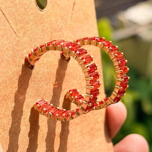 May include: Gold-toned hoop earrings encrusted with vibrant red rectangular gemstones. The earrings are displayed on a brown card, showcasing their design and color. The gemstones are evenly spaced, creating a continuous sparkle.