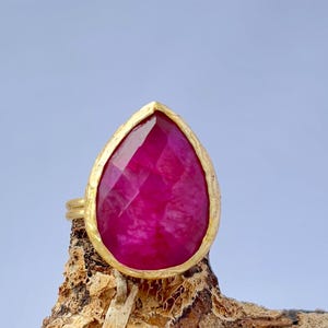 Peut inclure: Une bague dorée ornée d'une grande pierre précieuse en forme de goutte d'eau, facettée, de couleur rubis. Le rubis est d'un rouge-rosé vif. La bague est posée sur une surface en bois brun texturé, sur fond bleu clair.