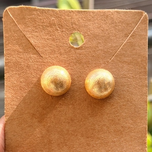 May include: Gold-coloured stud earrings with a textured, spherical design. The earrings are presented on a brown cardboard earring card. The background is blurred, with green foliage visible.