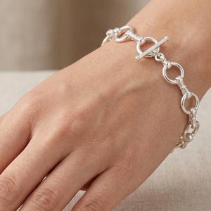 May include: A silver-toned chain bracelet featuring a series of circular links connected by small bars, secured with a T-bar toggle clasp. The bracelet has a polished metallic finish and a consistent link design throughout the entire length of the piece.