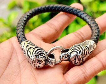 May include: A braided black leather bracelet featuring two polished silver-tone metal tiger head terminals that meet in the center, connected by a small circular ring clasp. The detailed tiger heads show engraved facial features.