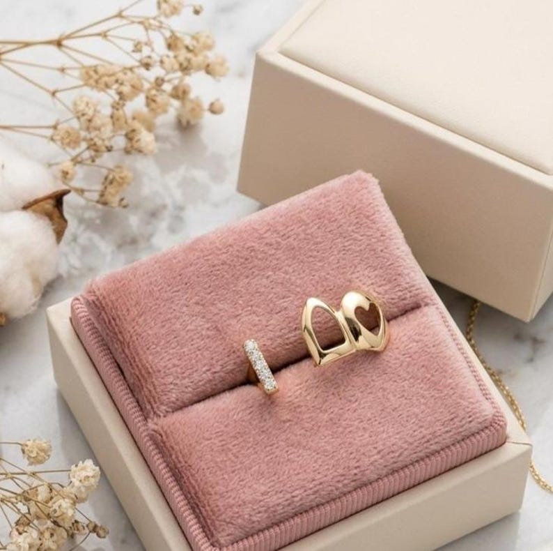 May include: A gold ring featuring a small, rectangular, diamond-encrusted bar and a larger, open-work gold element with a heart-shaped cutout, displayed on a soft, dusty pink velvet ring cushion inside a beige jewelry box.