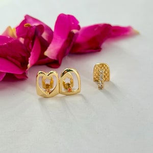 May include: Two gold-toned tooth grills. One piece features a heart-shaped cutout with a small star inside, while the other piece has a textured, grid-like surface with a vertical row of small, clear, sparkling stones set in the center.