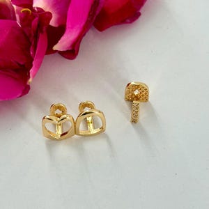 May include: A set of gold-toned metal tooth grills, including one piece shaped like a heart outline and another piece featuring a small, rectangular, textured surface with a vertical bar, both designed to fit over individual teeth.