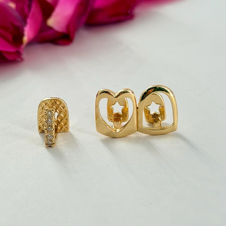 May include: A set of gold-toned tooth grills featuring a single piece with a vertical row of small clear stones and a double piece with cut-out heart and star shapes. The metal has a polished finish and a textured grid pattern on the single piece.