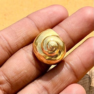 Könnte beinhalten: Ein kleines, poliertes, goldfarbenes Objekt in Form einer spiralförmigen Muschel. Die Oberfläche ist glatt und reflektierend und weist ein ausgeprägtes gewundenes Muster auf, das von der Mitte nach außen verläuft und eine runde, dreidimensionale Form bildet.