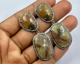 May include: A pair of drop earrings featuring three oval-shaped, faceted, multi-colored gemstones in shades of brown, tan, and red. Each gemstone is set in a dark metal frame bordered by small, clear, sparkling pavé-set stones.