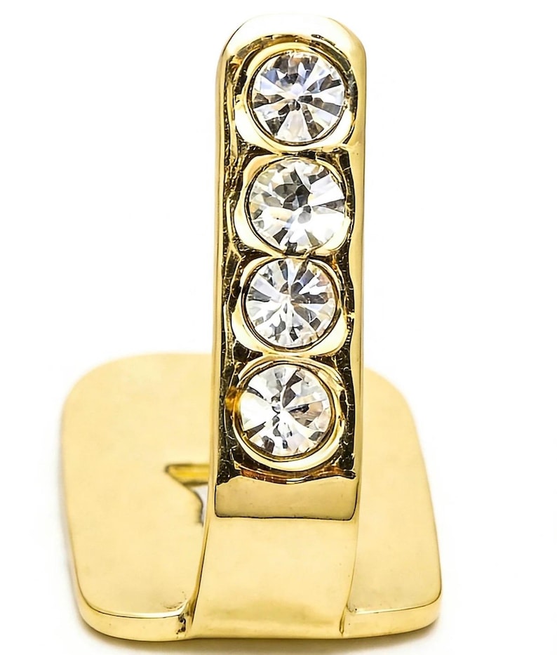 May include: A gold-toned metal ring featuring a vertical bar design adorned with four clear, round, faceted crystals set in a row. The band has a flat, rectangular base section that extends upward into the crystal-encrusted vertical element.