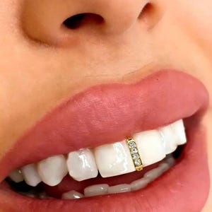 May include: A gold-toned tooth gem or grillz accessory featuring a vertical bar design set with three small, clear, round-cut stones. The piece is securely attached to the center of a single upper front tooth, positioned between the two central incisors.
